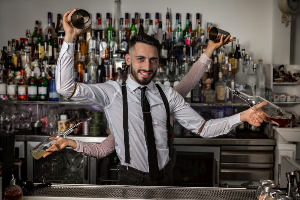 Cheerful Hispanic barman with hands of colleague behind back holding bottles of alcohol and shakers and looking at camera during work in bar. Bartenders with shakers and bottles in pub