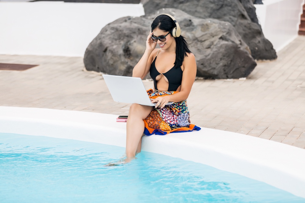 Young woman in bikini with headphones and sunglasses using laptop computer sitting on the edge of the pool enjoying her vacation