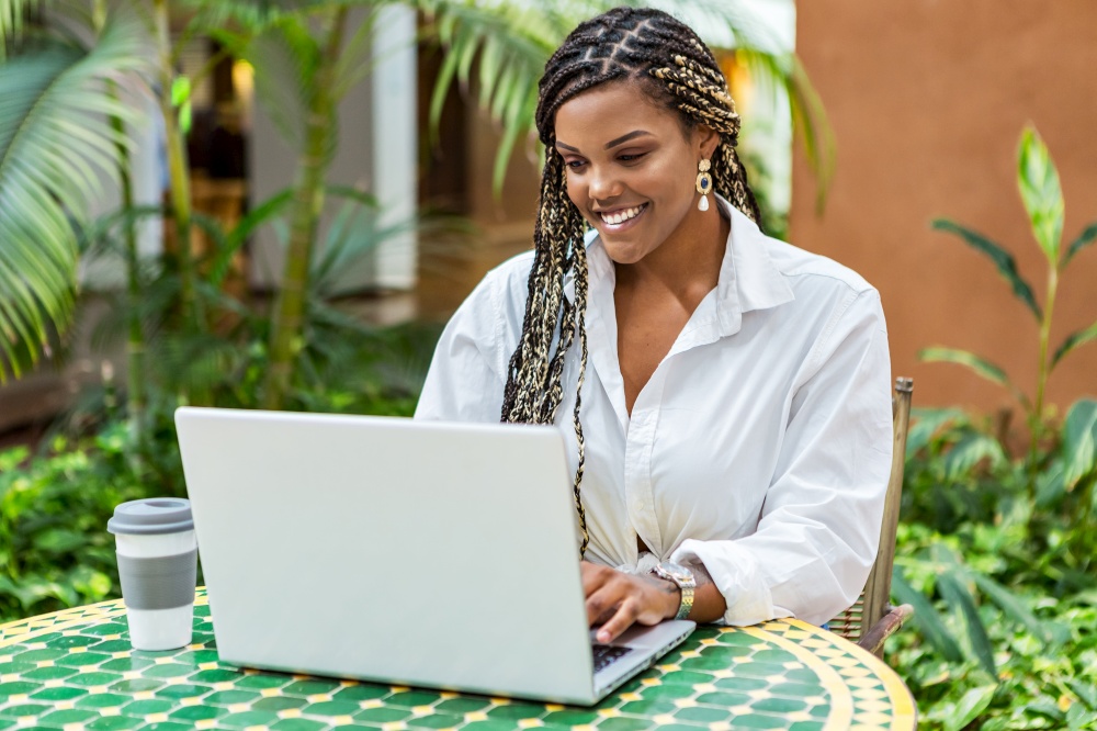 African American woman using laptop in a cafe store outside. Woman with braids sitting using laptop and drinking coffee.. African American woman using laptop in a cafe store outside