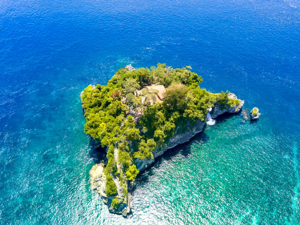 Indonesia. Emerald ocean water. A small rocky island, overgrown with jungles. Stairs from the water and a few huts between the trees. Aerial view. Small Rocky Island in the Ocean and Huts. Aerial View
