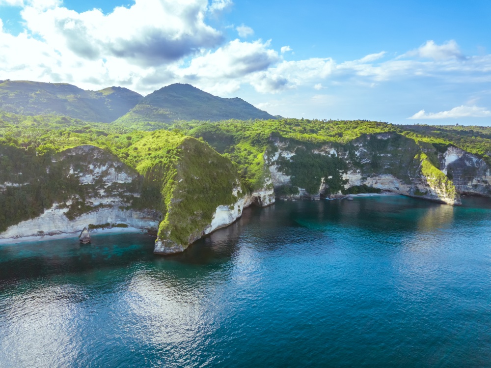 Indonesia. A large tropical island overgrown with jungle. Rocky shore of the ocean. The clouds on the blue sky. Aerial view. Rocky Shore of the Tropical Island and Clouds. Aerial View