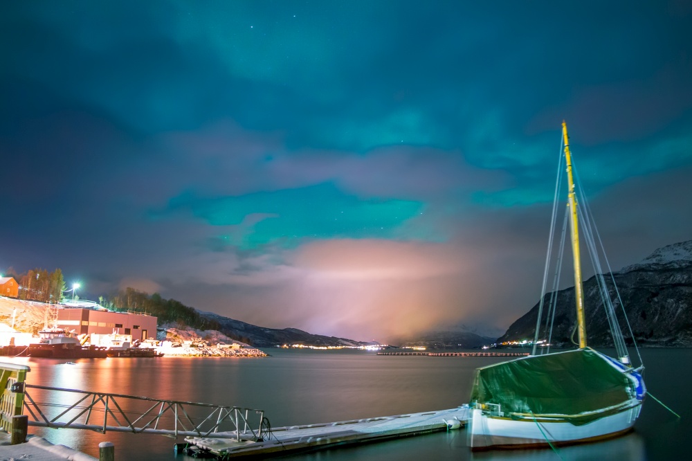 Night fjord in winter Norway. The boat is at the pier. The lights of the village on the horizon, surrounded by snow-capped mountains. Small northern lights in the sky. Boat and Village in Winter Norway at Night