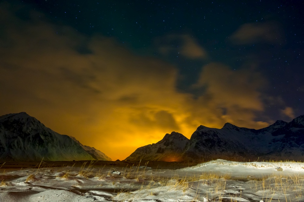 Winter Norway. Grass covered with snow in the night valley. The lights of the city highlight the clouds behind the mountains. Snow-Covered Grass and Glow beyond the Mountains