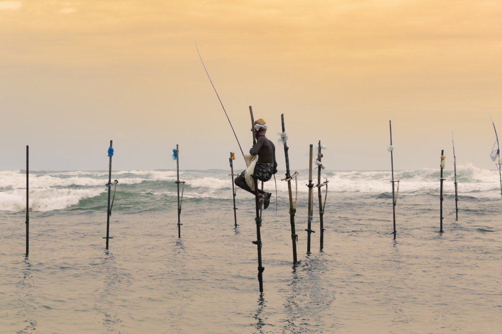 Stilt fisherman sitting in his pole with a wooden fishing rod in his hands in the sunset evening. Ocean waves crash behind him in the background.