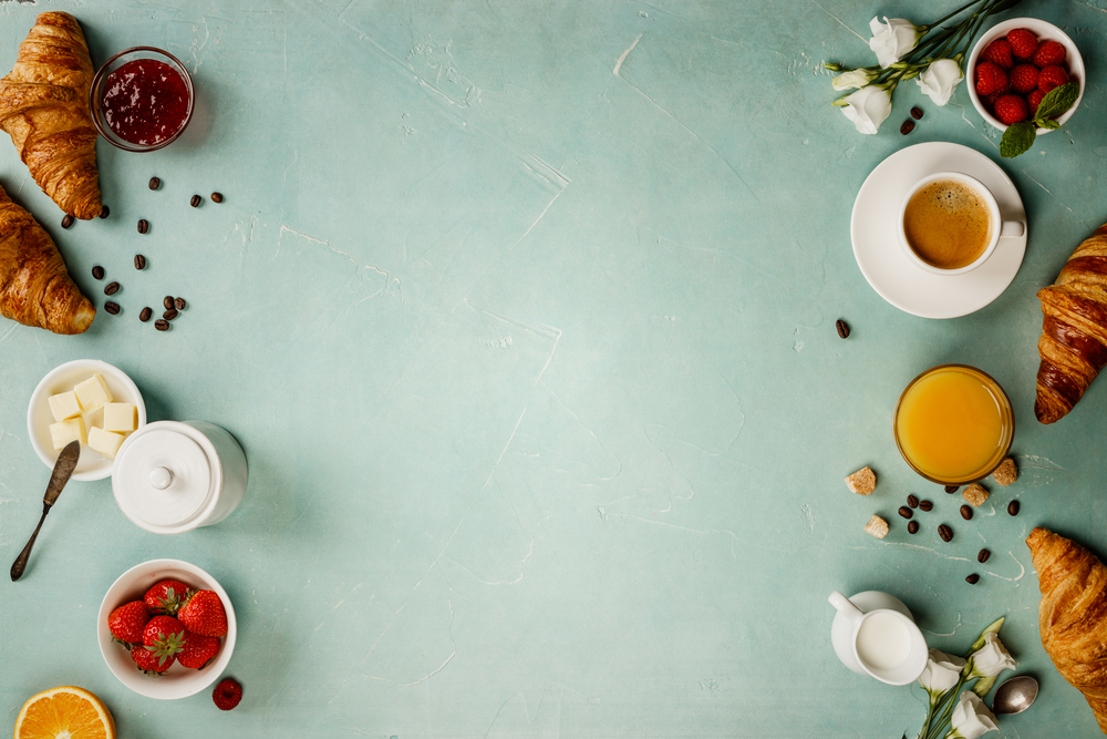 Continental breakfast captured from above top view, flat lay . Coffee, orange juice, croissants, jam, berry, milk and flowers. Blue concrete worktop as background. Layout with free text copy space.. Continental breakfast captured from above - space for text