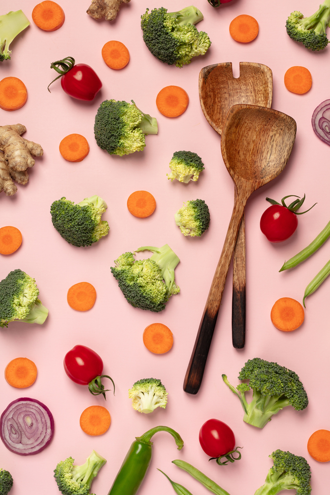 Colorful pattern of tomatoes, broccoli, carrots, ginger and onion with wooden salad spoons on a pink background. Top view of sliced seasonal vegetables. The concept of a healthy diet