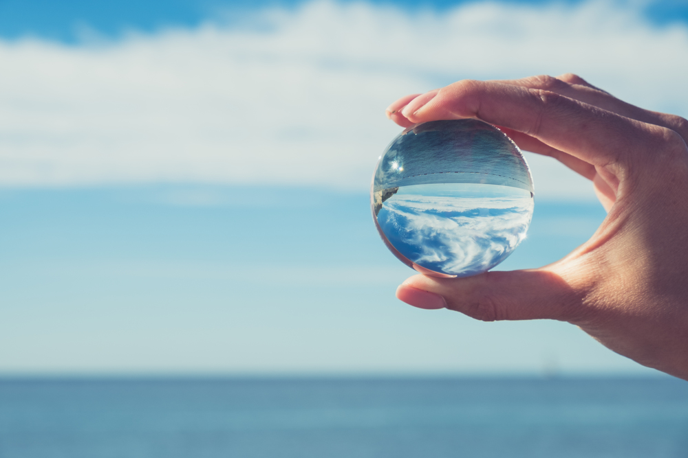 Woman&rsquo;s hand holding a crystal ball, looking through to the ocean and sky. Creative photography, crystal ball refraction