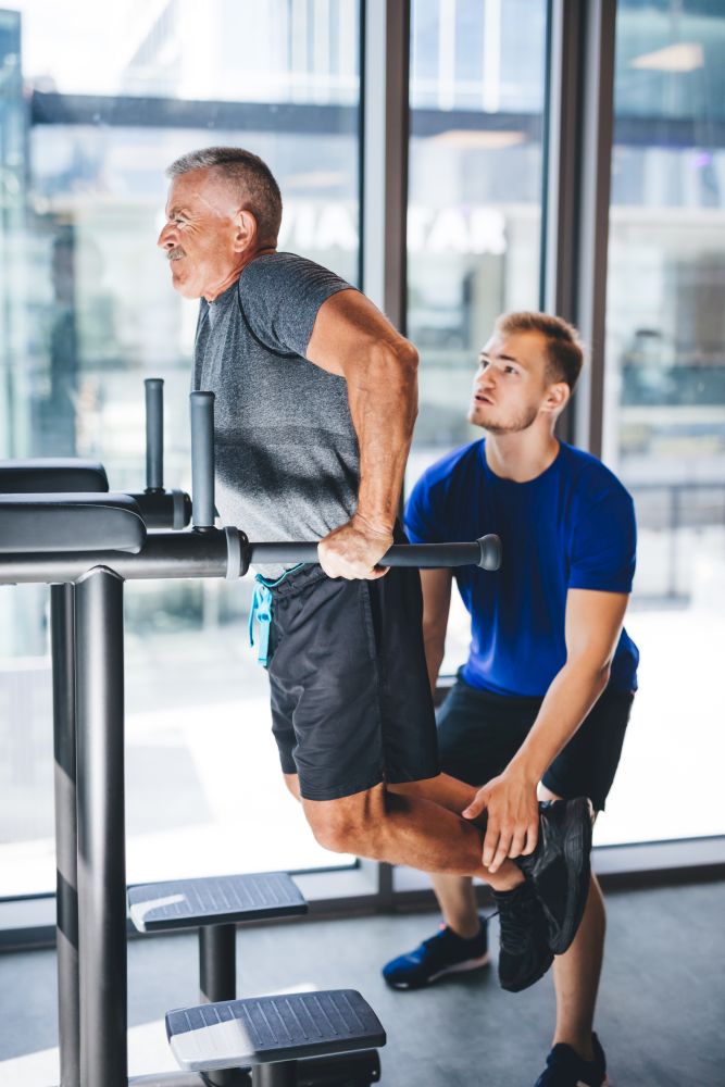 Young man helping senior man with his workout at a gym. Personal trainer. Active retirement.. Man helping senior man with his workout at a gym.