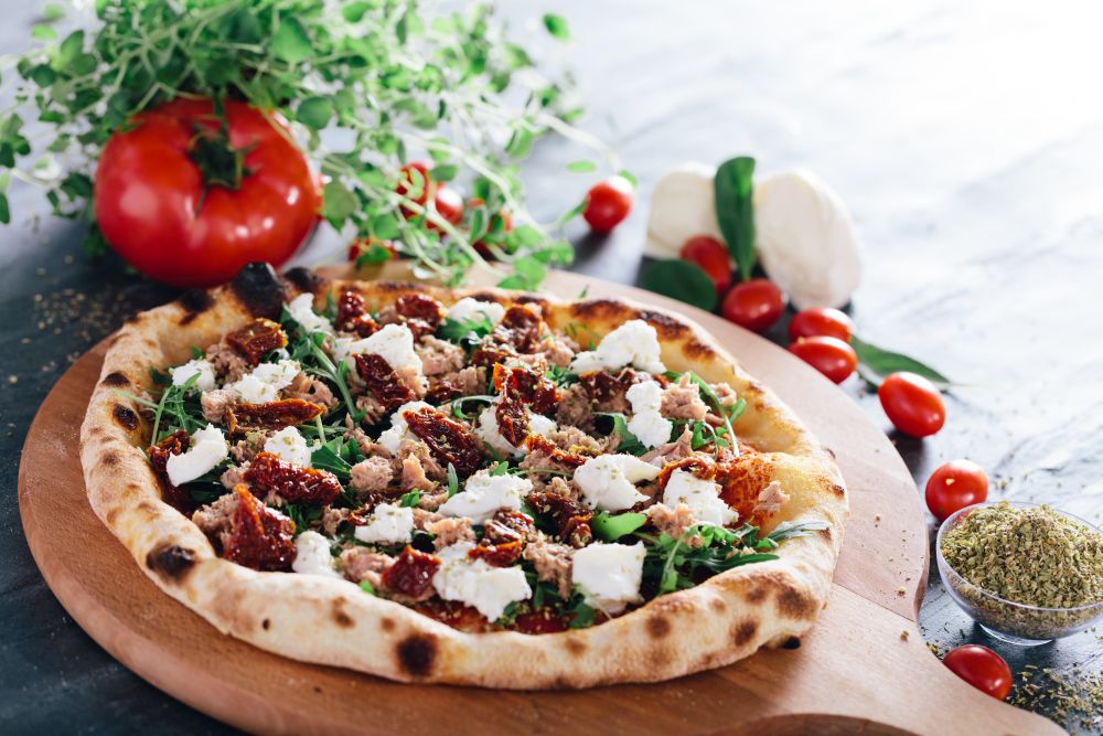 Pizza with dried tomatoes, mozzarella, sausage and rocket salad on wooden board. Oregano leaves, tomatoes and mozzarella in the background. Popular traditional food.. Pizza with dried tomatoes, mozzarella, sausage and rocket