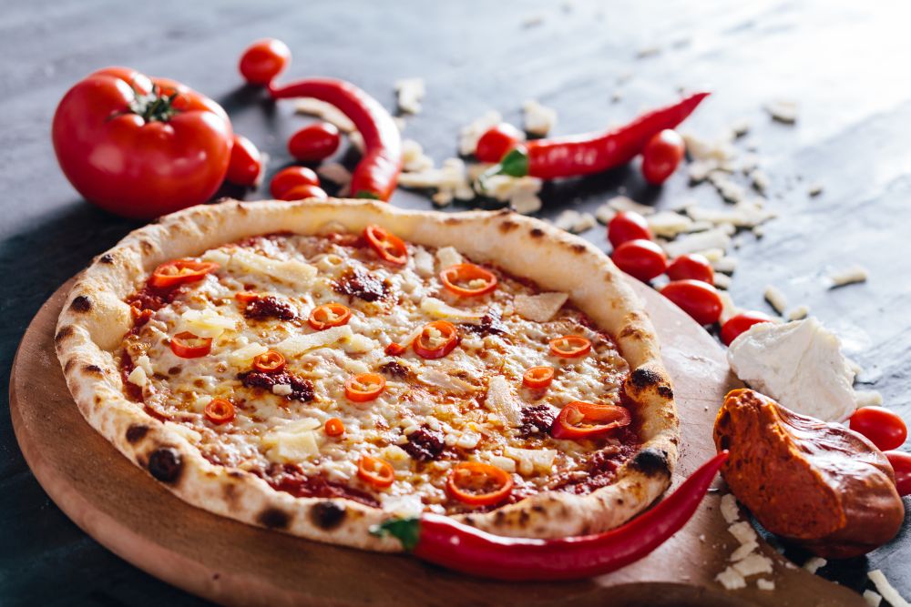 Pizza with hot pepper, cheese and meat spread on wooden board. Tomatoes, hot peppers, parmesan and mozzarella in the background. Popular traditional food.. Pizza with hot pepper, cheese and meat spread