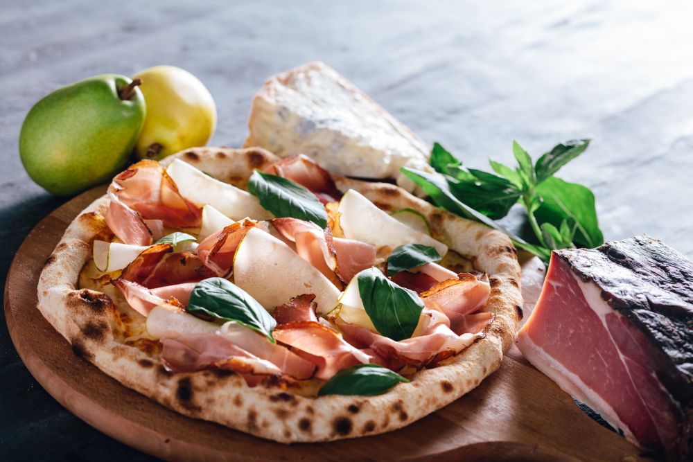Pizza with ham, pear, basil and blue cheese on wooden board. Ham, pear, blue cheese and basil leaves in the background. Popular traditional food.. Pizza with ham, pear, basil and blue cheese