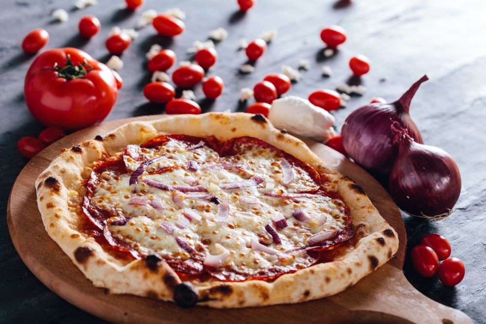Pizza with salami, mozzarella and red onion on wooden board. Tomatoes, mozzarella and red onions in the background. Popular traditional food.. Pizza with salami, mozzarella and red onion on wooden board.