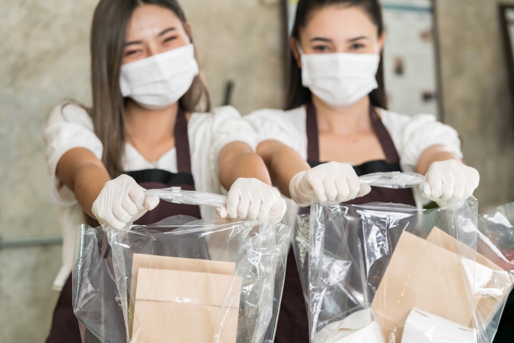 Close up waitress wear protective face mask smile and hold shopping bag for take away or takeout food. This essential service is very popular while city lockdown from coronavirus COVID-19 Pandemic