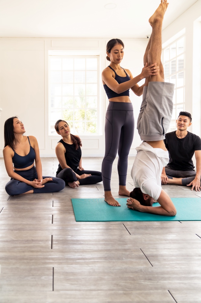 Asian people learning Yoga class in fitness club. Instructor coaching and adjust correct head stand pose to student at front while others looking. Yoga Practice Work out lifestyle concept.