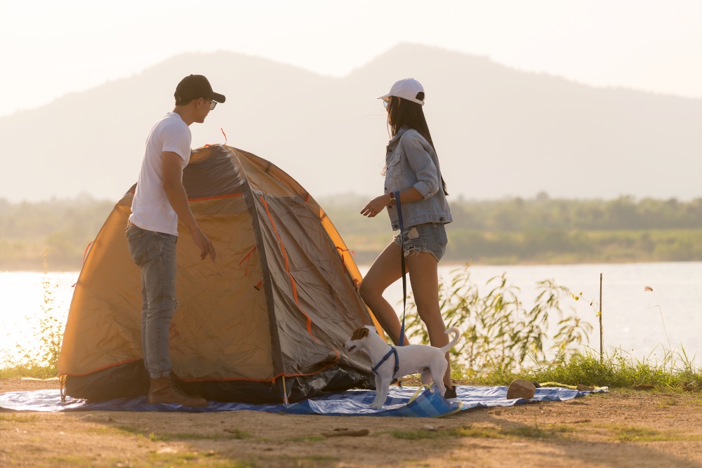 Young adult Asian couple pitch and set up tent for camping around the lake. Camping trip with dog outdoor activity campsite concept.