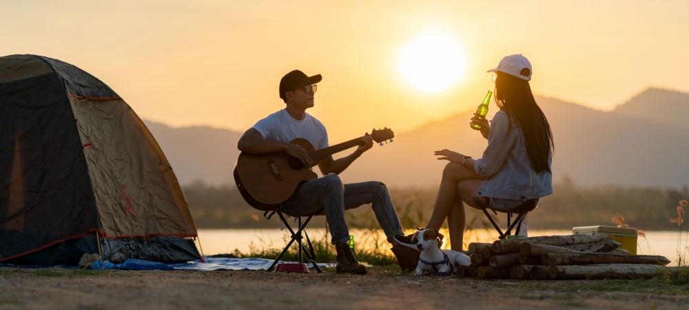 Panoramic Young adult Asian couple playing guitar and drinking beer beside their tent campsite while sunset. Camping trip with dog outdoor activity campsite concept.