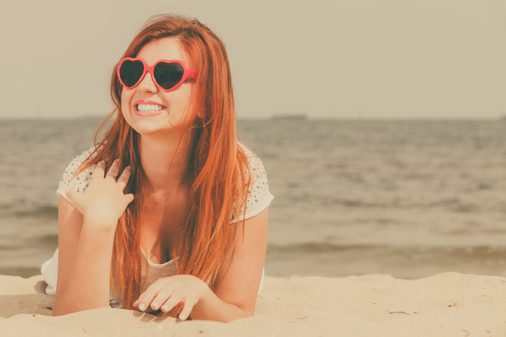 Summer relaxation and recreation concept. Happy redhead adult woman wearing sunglasses in heart shape lying on beach during summertime.. Redhead adult woman lying on beach