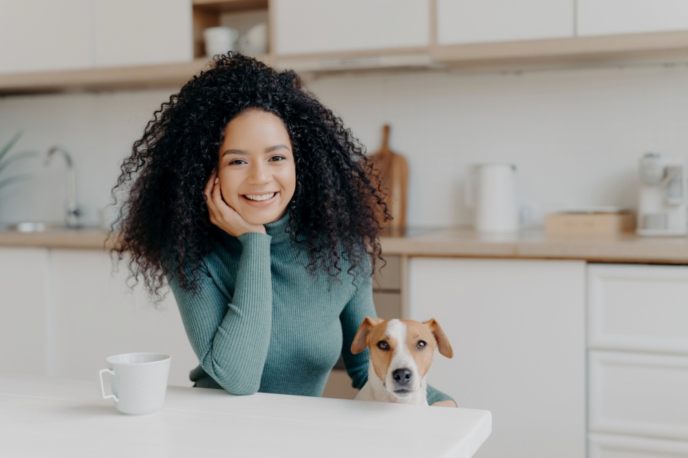 Joyful woman with curly hair and her dog at a kitchen table, sharing a moment of happiness