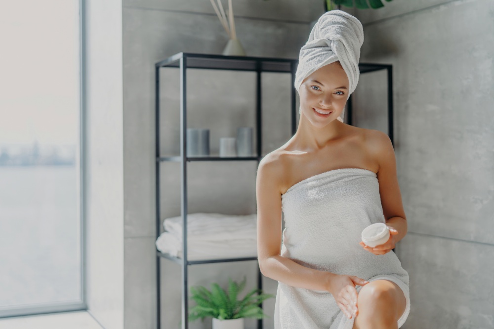 A woman with a towel on her head smiles, holding a skincare product in a minimalist spa