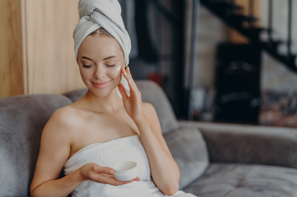 A woman with a towel headwrap applies face cream, seated comfortably on a couch