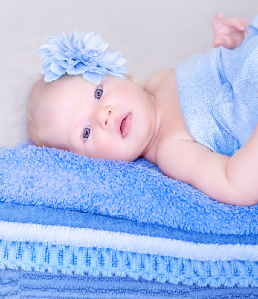 Closeup portrait of sweet newborn girl lying down on many soft blue towels, cute flower on head, innocence and tenderness concept