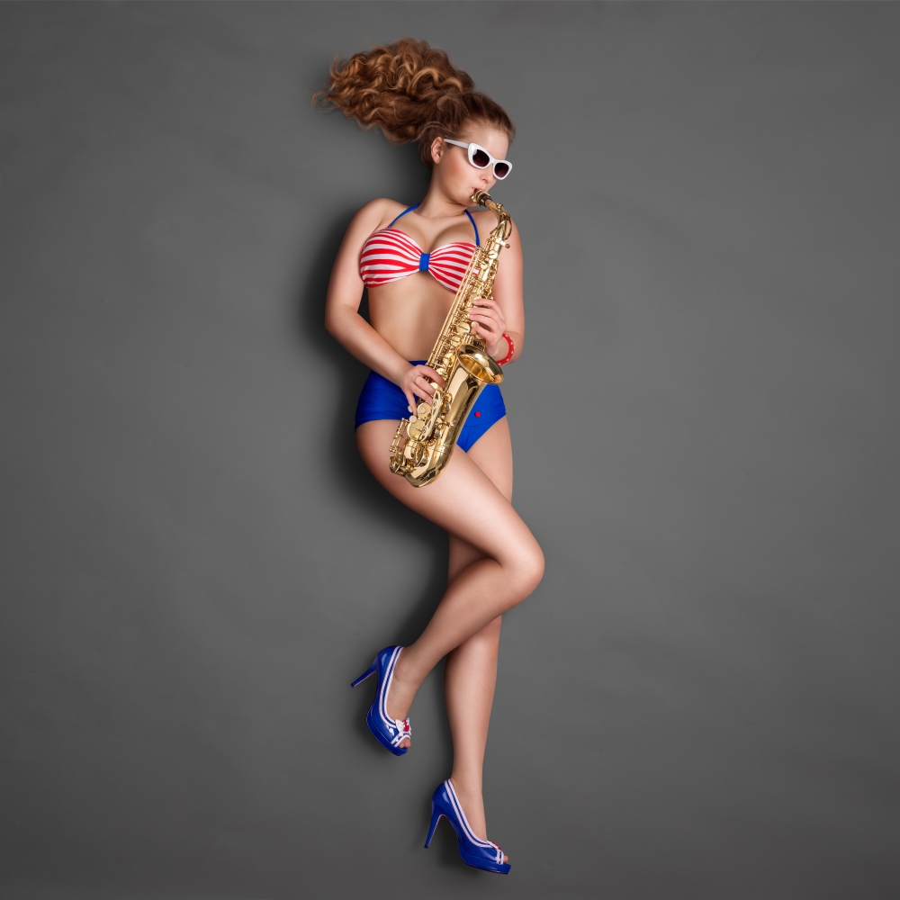 Top view of a beautiful pin-up girl in retro bikini and sunglasses, playing jazz saxophone on chalkboard background.