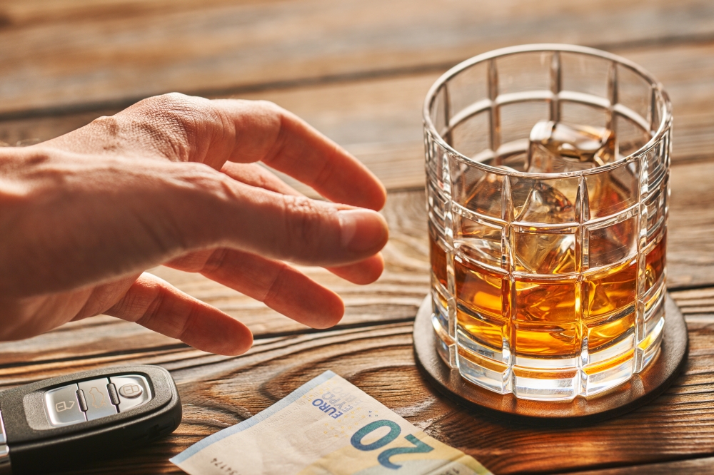 Man's hand reaching to glass of whiskey or alcohol drink with ice cubes and car key on rustic wooden table. Drink and drive and alcoholism concept. Safe and responsible driving concept.