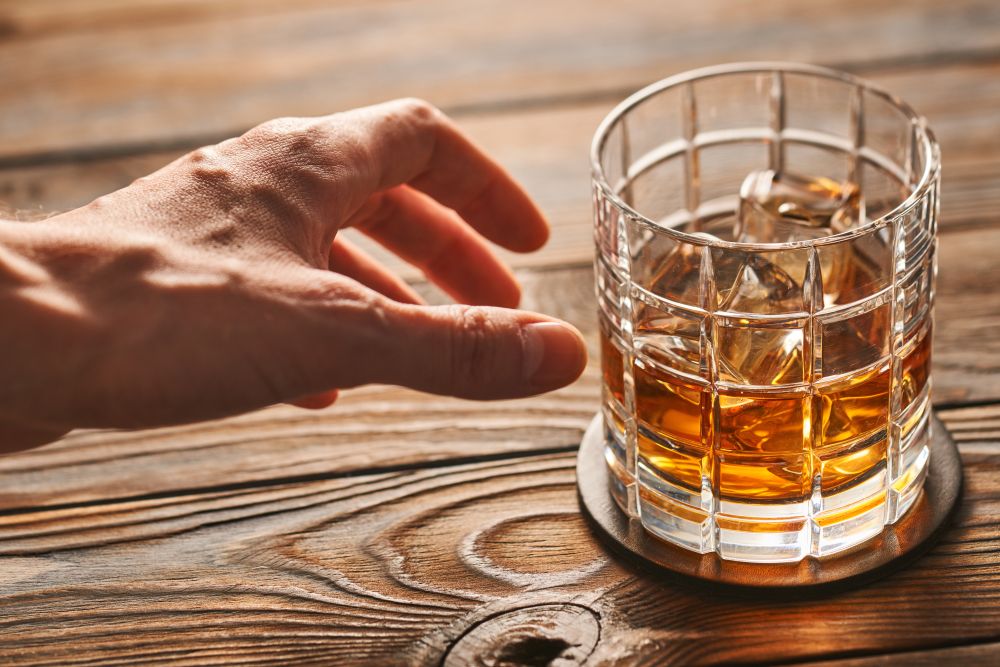 Man&rsquo;s hand reaching to glass of whiskey with ice cubes on rustic wooden table. Alcoholism concept.