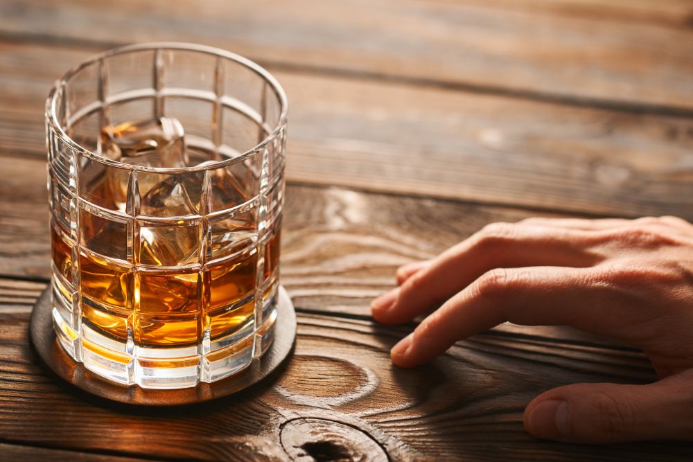 Glass of whiskey with ice cubes and man&rsquo;s hand on rustic wooden table with copy-space. Alcoholism concept.