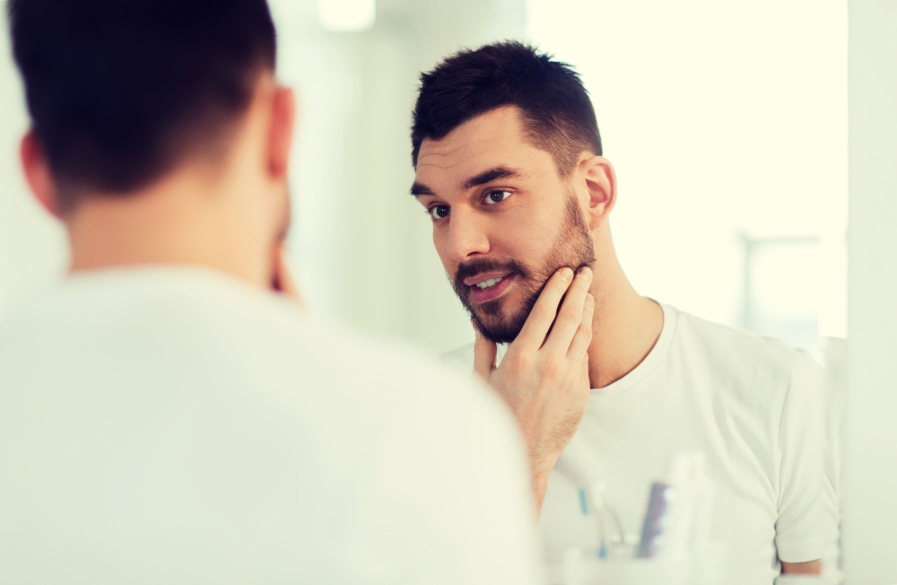 beauty, hygiene and people concept - smiling young man looking to mirror at home bathroom. happy young man looking to mirror at home bathroom