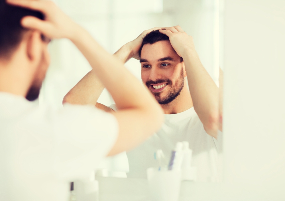 beauty, hygiene and people concept - smiling young man looking to mirror and styling hair at home bathroom. happy young man looking to mirror at home bathroom