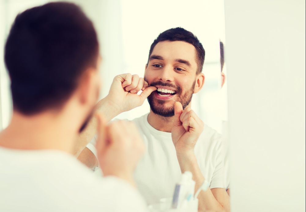 health care, dental hygiene, people and beauty concept - smiling young man with floss cleaning teeth and looking to mirror at home bathroom. man with dental floss cleaning teeth at bathroom