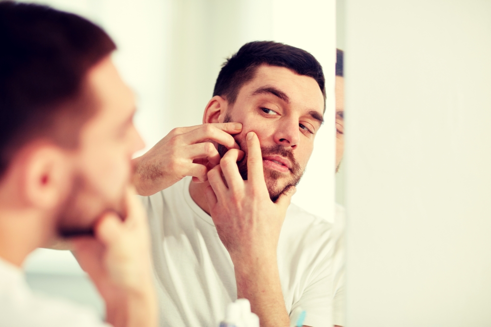 beauty, hygiene, skin problem and people concept - young man looking to mirror and squeezing pimple at home bathroom. man squeezing pimple at bathroom mirror