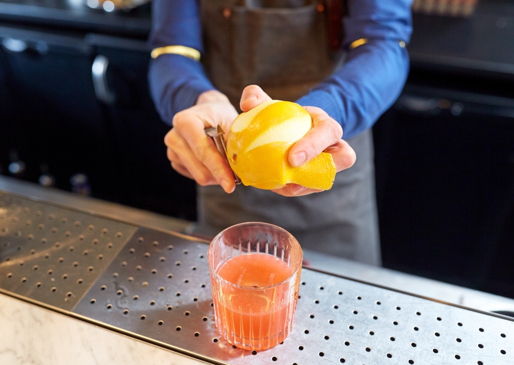 alcohol drinks, people and luxury concept - bartender with glass and peeler removing peel from lemon and preparing cocktail at bar. bartender removing peel from lime at bar