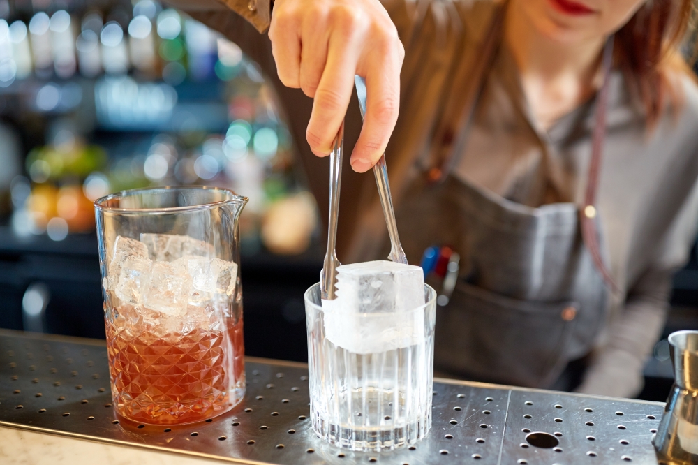 alcohol drinks, people and luxury concept - woman bartender with tongs adding ice cube into glass and preparing cocktail at bar counter. bartender adding ice cube into glass at bar