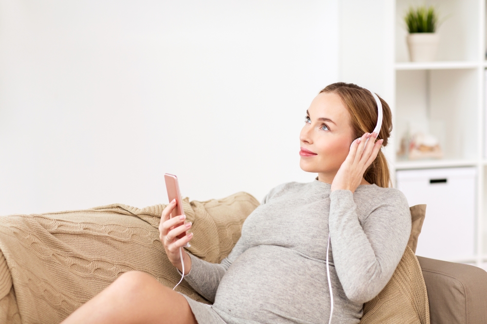 pregnancy, motherhood, technology, people and expectation concept - happy pregnant woman with smartphone and headphones listening to music at home. pregnant woman with smartphone and headphones