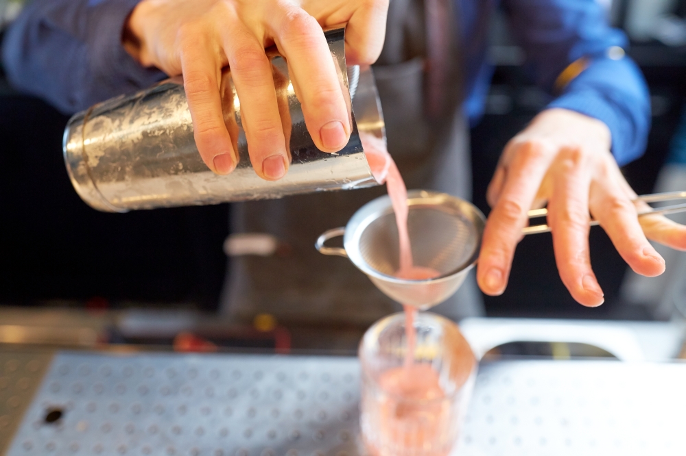 alcohol drinks, people and luxury concept - barman pouring cocktail from shaker through strainer into glass at bar. barman with shaker preparing cocktail at bar