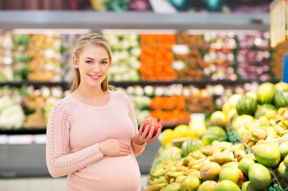 sale, shopping, food, pregnancy and people concept - happy pregnant woman with pomegranate at grocery store or supermarket. happy pregnant woman with pomegranate at grocery