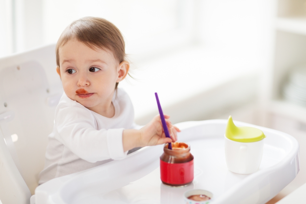 food, child, feeding and people concept - baby with spoon sitting in highchair and eating puree from jar at home kitchen. baby with spoon eating puree from jar at home