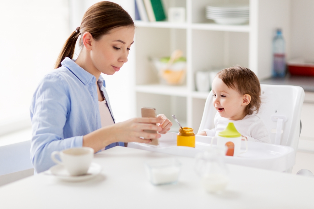family, breakfast, eating, technology and people concept - happy mother with smartphone and coffee and little baby sitting in highchair at home kitchen. mother with smartphone and baby eating at home