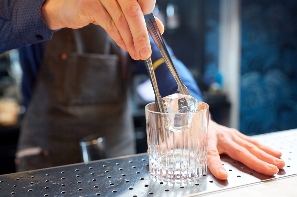 alcohol drinks, people and luxury concept - bartender with tongs adding ice cube into glass and preparing cocktail at bar counter. bartender adding ice cube into glass at bar
