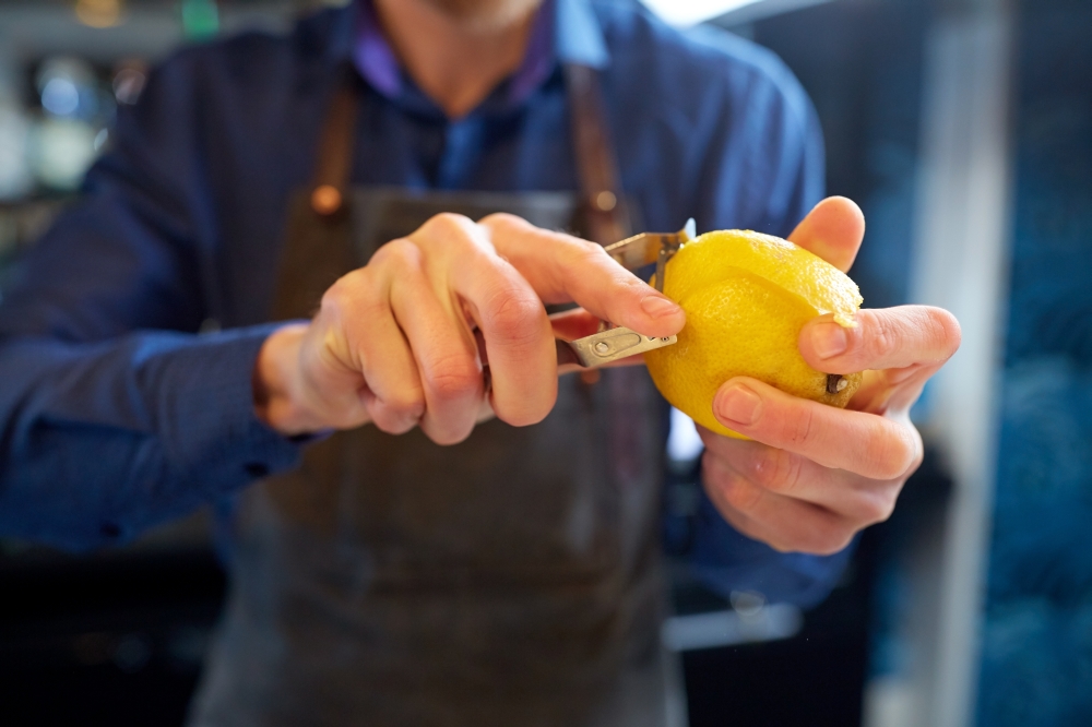 people and profession concept - bartender with peeler removing peel from lemon at bar. bartender removing peel from lime at bar