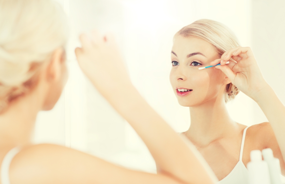 beauty, hygiene and people concept - smiling young woman fixing makeup with cotton swab and looking to mirror at home bathroom. woman fixing makeup with cotton swab at bathroom