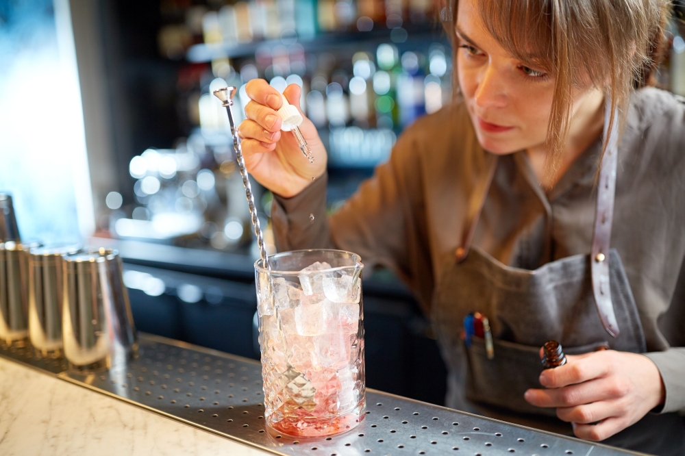 alcohol drinks, people and luxury concept - woman bartender preparing cocktail and adding essence into glass with ice at bar counter. bartender adding essence to cocktail glass at bar