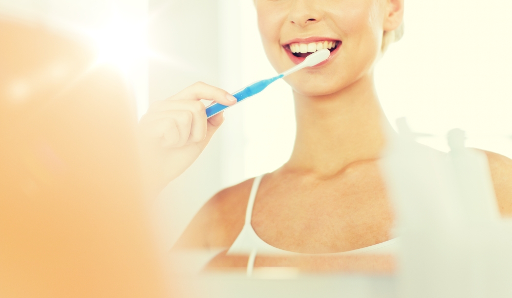 health care, dental hygiene, people and beauty concept - close up of smiling young woman with toothbrush cleaning teeth and looking to mirror at home bathroom. woman with toothbrush cleaning teeth at bathroom. woman with toothbrush cleaning teeth at bathroom