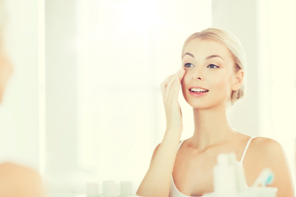 beauty, skin care and people concept - smiling young woman applying cream to face and looking to mirror at home bathroom. happy woman applying cream to face at bathroom. happy woman applying cream to face at bathroom