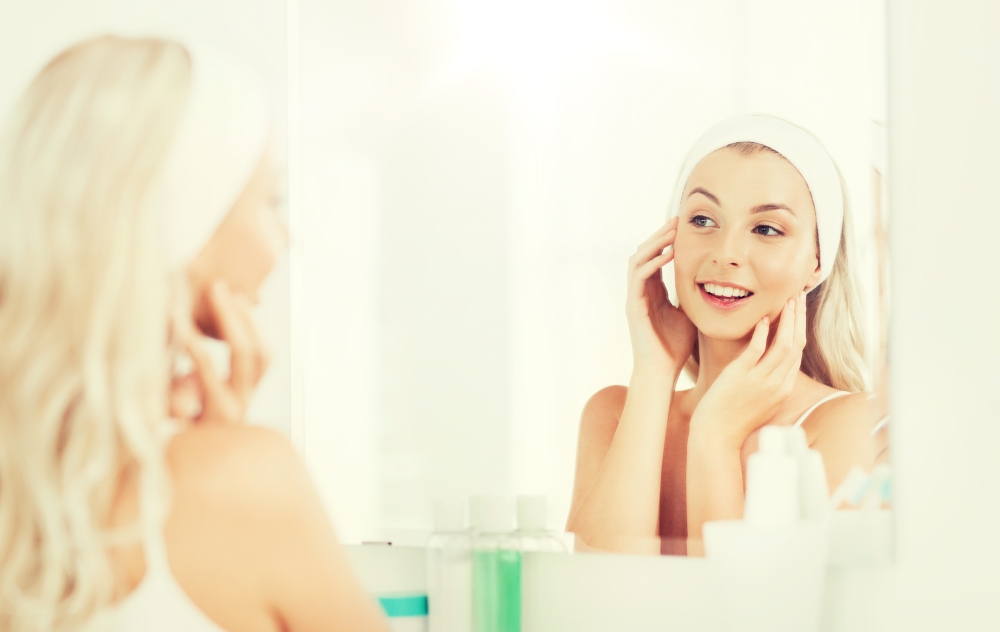 beauty, skin care and people concept - smiling young woman in hairband touching her face and looking to mirror at home bathroom. woman in hairband touching her face at bathroom. woman in hairband touching her face at bathroom