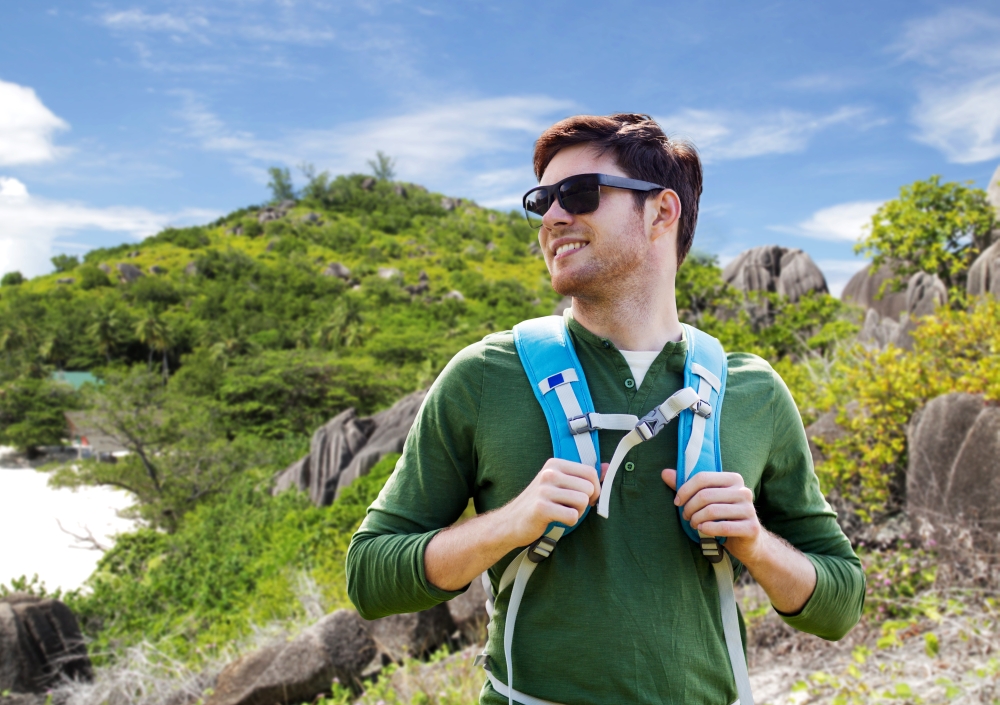 travel, tourism and people concept - happy young man in sunglasses with backpack over natural exotic island background. happy man with backpack traveling around island. happy man with backpack traveling around island