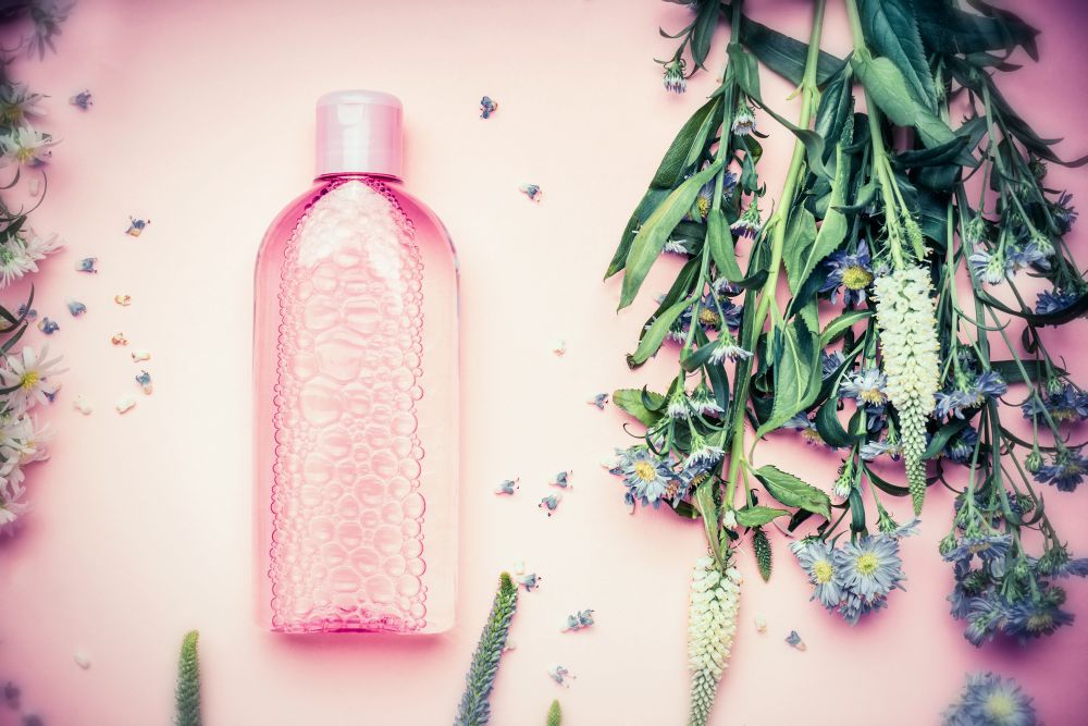 Plastic bottle with tonic or micellar cleansing water with fresh herbs and flowers on pink background, top view.  Beauty, skin, hair or body care concept