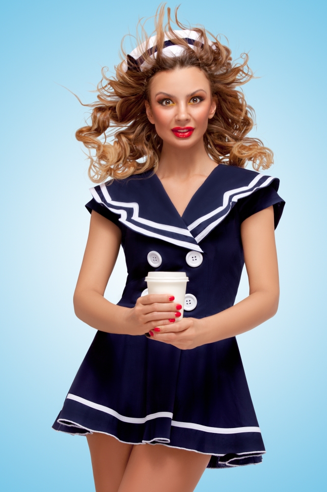 Creative photo of a playful pin-up sailor girl with a coffee-to-go cup, having wind in her hair on blue background.